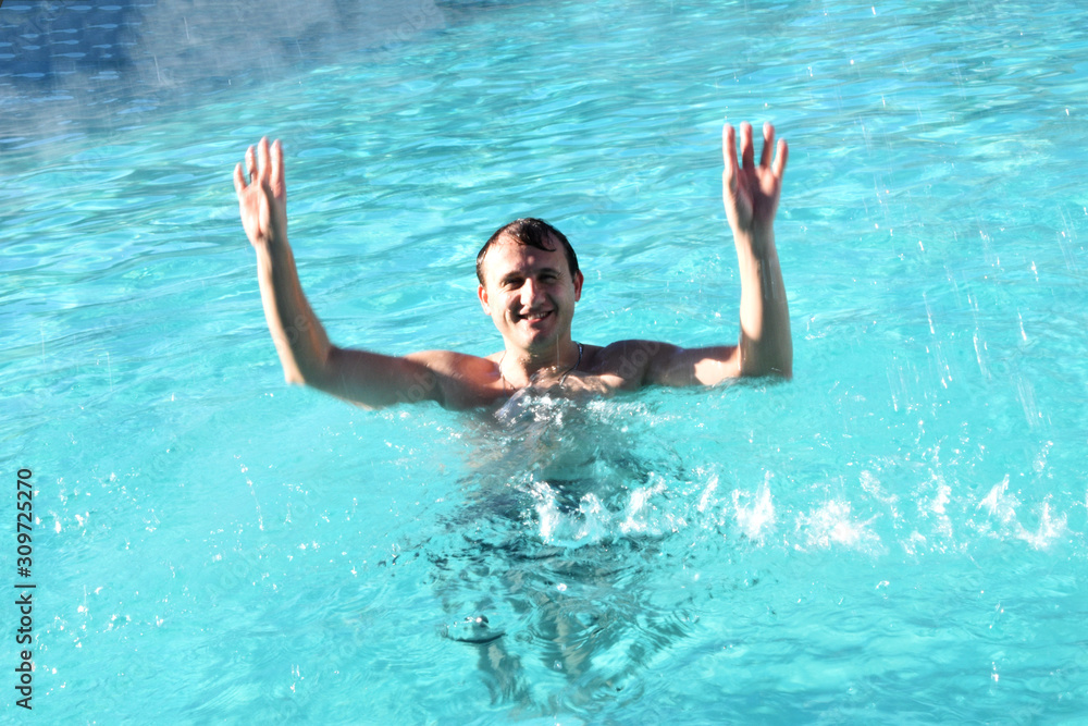 A young man in the pool has fun, around a lot of water spray. The turquoise, clear water in the pool is fun and funny to swim.