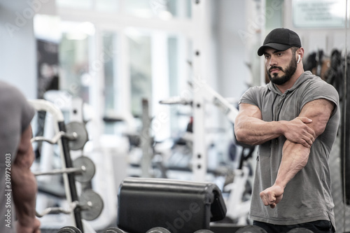 strong handsome young caucasian bearded man in cap and sportswear with wireless headphones holding hand painful elbow joint stretching muscle injury in fitness gym