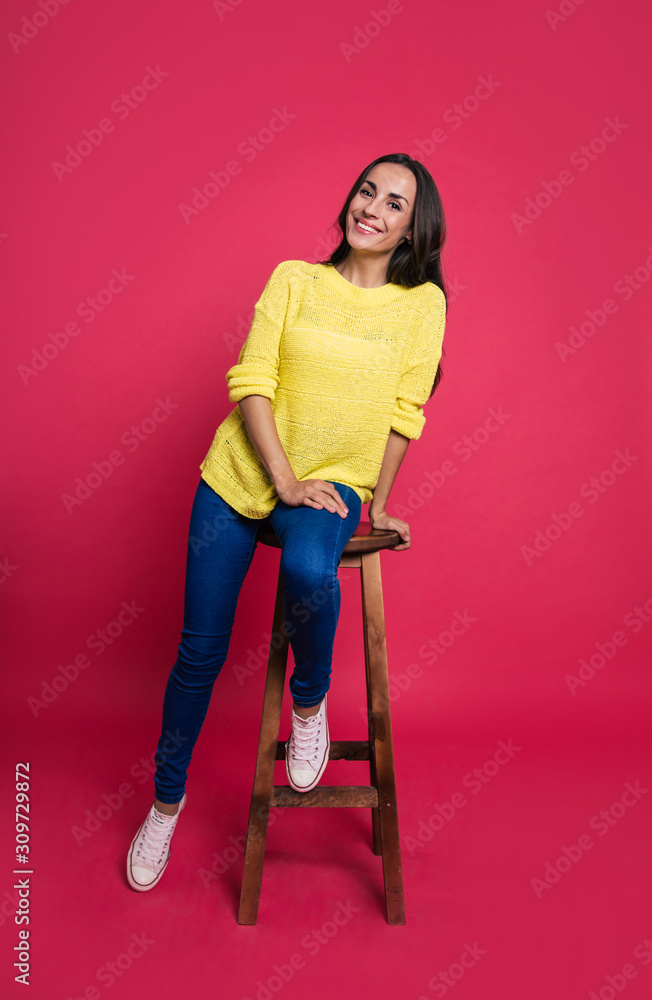 Sunny colors. Full-length photo of a beautiful girl in a yellow sweater, who is sitting on a wooden chair, smiling and looking right in the camera.