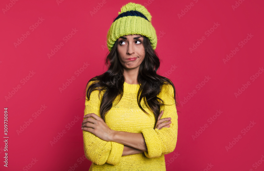 Skeptical look. Fascinating woman in a bright yellow sweater and a green hat with pompom is posing in front with her arms folded, looking to the left with a skeptical facial expression. Skeptical look. Fascinating woman in a bright yellow sweater and a green hat with pompom is posing in front with her arms folded, looking to the left with a skeptical facial expression.