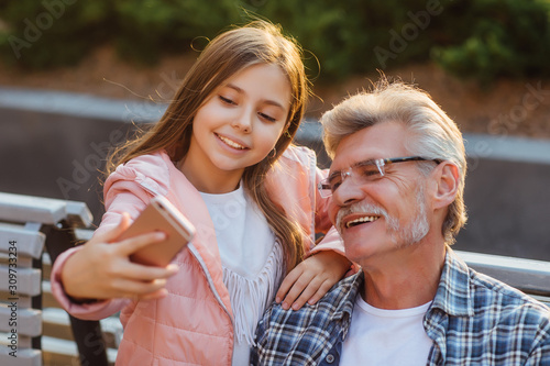 Photo of a grandfather and granddaughter sitting on a bench and speak online by smartphone.
