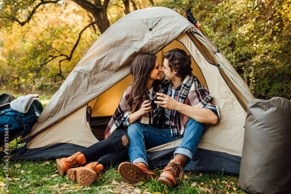 Young beautiful couple in casual dress sitting in a campsite near the tent.  Attractive woman and handsome boyfriend have fun, drink tea and sitting on  plaid in nature. Stock Photo | Adobe