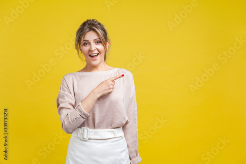 Look attention! Portrait of surprised beautiful young woman with fair hair in casual blouse standing, pointing aside, looking excited and shocked. indoor studio shot isolated on yellow background