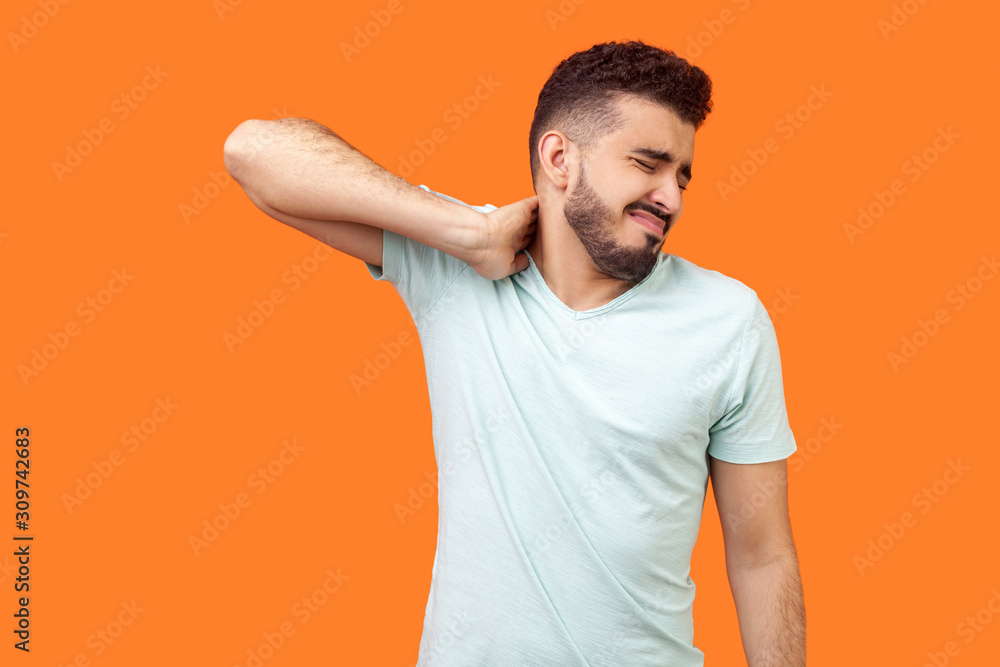 Portrait of exhausted brunette man with beard in white t-shirt rubbing his neck and grimacing from pain, suffering muscle tension, shoulder hurts. indoor studio shot isolated on orange background Portrait of exhausted brunette man with beard in white t-shirt rubbing his neck and grimacing from pain, suffering muscle tension, shoulder hurts. indoor studio shot isolated on orange background