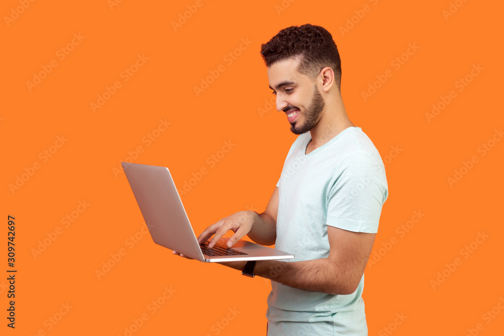 Side view portrait of cheerful brunette man with beard in white t-shirt working on laptop typing email or chatting in social network, happy face expression. studio shot isolated on orange background Side view portrait of cheerful brunette man with beard in white t-shirt working on laptop typing email or chatting in social network, happy face expression. studio shot isolated on orange background