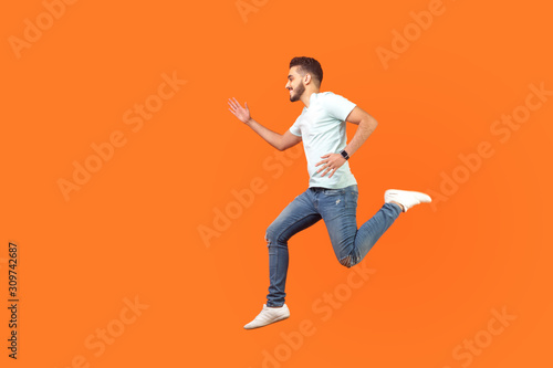 Side view of joyous brunette man with beard in sneakers and denim outfit running in air, hurrying for discounts, empty copy space for advertising. indoor studio shot isolated on orange background
