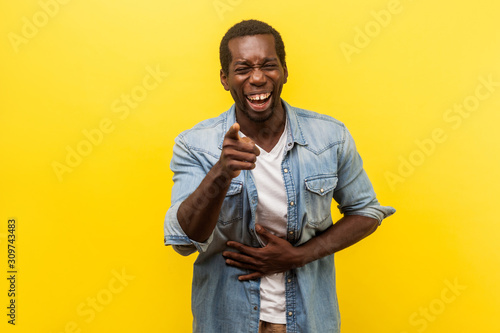 Portrait of joyful positive man in denim shirt with rolled up sleeves holding hand on belly and laughing out loud, pointing at camera, mocking you. indoor studio shot isolated on yellow background