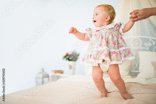 Moms hand holds a charming little girl in a dress taking her first steps on a chic double bed against a blue wall and decor. Concept baby products