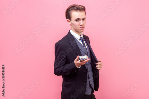 Portrait of arrogant rich businessman in elegant suit and with stylish haircut showing bunch of dollars on camera, offering money or boasting earnings. indoor studio shot isolated, pink background