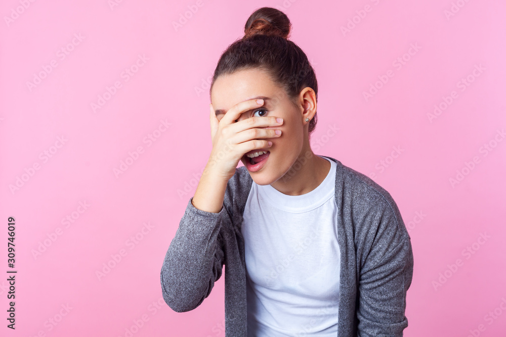 Portrait of adorable curious brunette teen girl with bun hairstyle in casual clothes peeking through fingers, looking embarrassed, pleasantly surprised. indoor studio shot isolated on pink background Portrait of adorable curious brunette teen girl with bun hairstyle in casual clothes peeking through fingers, looking embarrassed, pleasantly surprised. indoor studio shot isolated on pink background