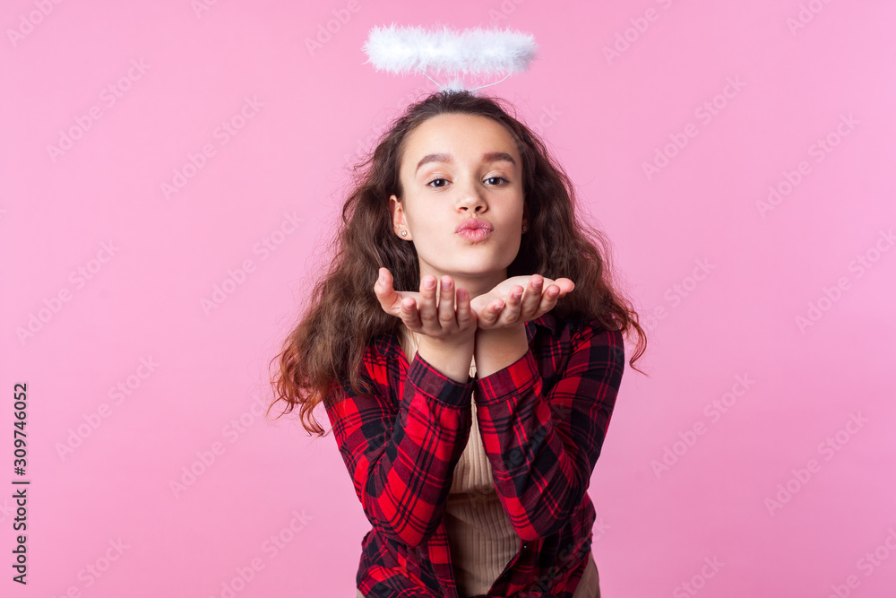 Portrait of lovely teenage girl with curly brunette hair in plaid shirt and toy halo above head sending air kiss with pouted lips, romance on valentines day. studio shot isolated on pink background Portrait of lovely teenage girl with curly brunette hair in plaid shirt and toy halo above head sending air kiss with pouted lips, romance on valentines day. studio shot isolated on pink background
