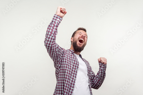 Hurray! Portrait of overjoyed winner, bearded man in casual plaid shirt raising hands, screaming yes i did it, emotionally reacting to success, victory. indoor studio shot isolated on white background photo
