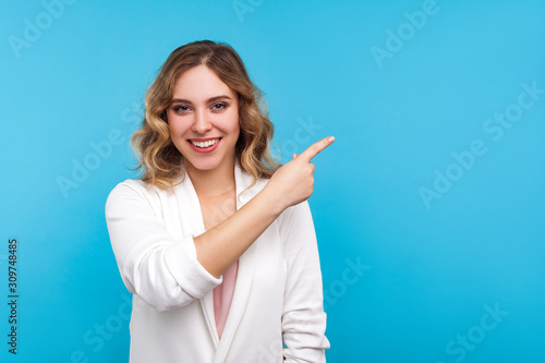 Look here  advertise  Portrait of positive woman with wavy hair in white jacket smiling happily and pointing to empty place for idea  copy space wall. indoor studio shot isolated on blue background