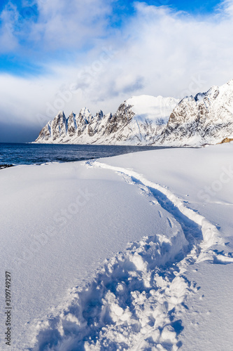 Snowy winter landscape of Lofoten Islands in Norway  scenery of island Senja - snow capped beach against of dramatic blue cloudy sky and jagged mountain ridge. Traditional Arctic Scandinavian view.