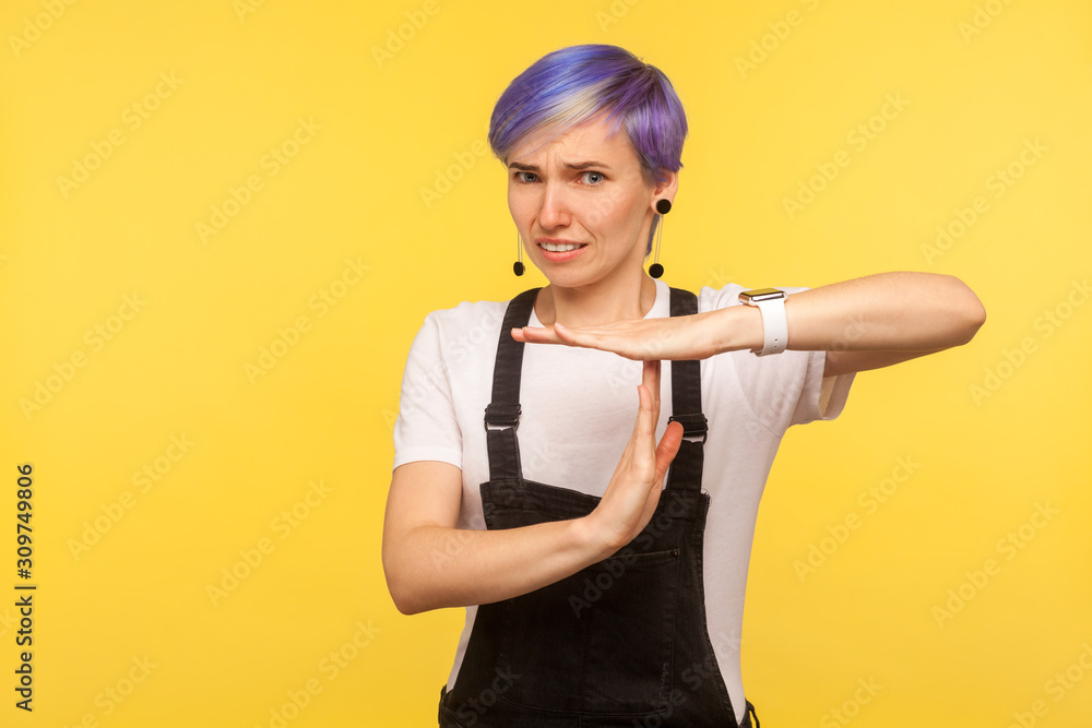 Portrait of tired hipster woman with violet short hair in denim overalls showing timeout gesture with hands and looking imploringly, worker need more time. isolated on yellow background, studio shot Portrait of tired hipster woman with violet short hair in denim overalls showing timeout gesture with hands and looking imploringly, worker need more time. isolated on yellow background, studio shot