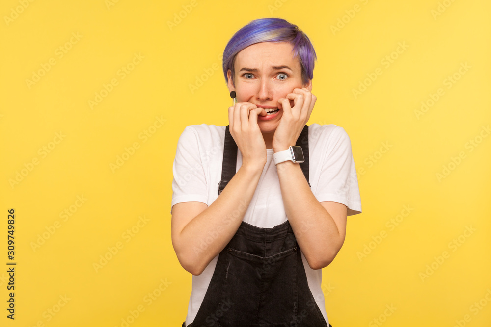Portrait of nervous worried hipster girl with violet short hair in denim overalls biting nails fingers, looking at camera with anxiety, feeling stressed out, nervous. yellow background, studio shot Portrait of nervous worried hipster girl with violet short hair in denim overalls biting nails fingers, looking at camera with anxiety, feeling stressed out, nervous. yellow background, studio shot
