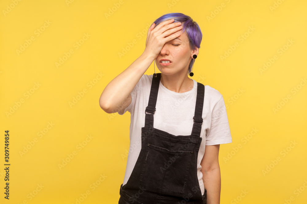 Facepalm. Portrait of forgetful hipster girl with violet short hair in denim overalls holding hand on forehead, expressing regret, sorrow, blaming herself for mistake. yellow background, studio shot Facepalm. Portrait of forgetful hipster girl with violet short hair in denim overalls holding hand on forehead, expressing regret, sorrow, blaming herself for mistake. yellow background, studio shot