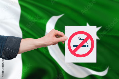 Pakistan health concept. Hand holding paper with no smoking sign over national waving flag. Quit smoke theme.