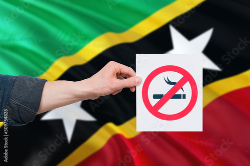 Saint Kitts And Nevis health concept. Hand holding paper with no smoking sign over national waving flag. Quit smoke theme.