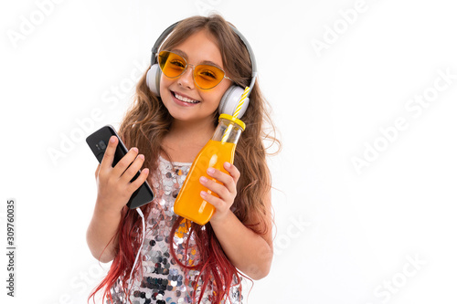 Little pretty caucasian girl listen to music with big earphones and drinks orange juice, picture isolated on white background