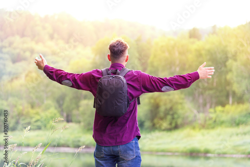 a man with a backpack, with arms spreading to the sides, stands by the river and looks at the bright sunny sky. concept of travel and freedom