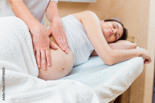 A beautiful pregnant brunette with long hair lies on her side at a reception with a massage therapist in a cosmetology room