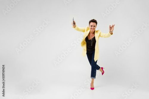 pretty brunette with a short haircut in a cream-colored jacket and pink pumps pumps dancing with a phone in her hands on a gray background