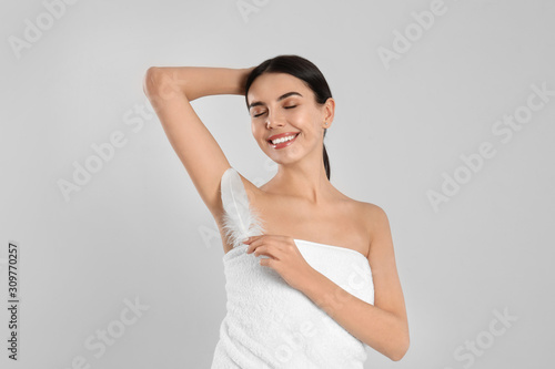 Young woman touching armpit with feather after epilation procedure on light grey background