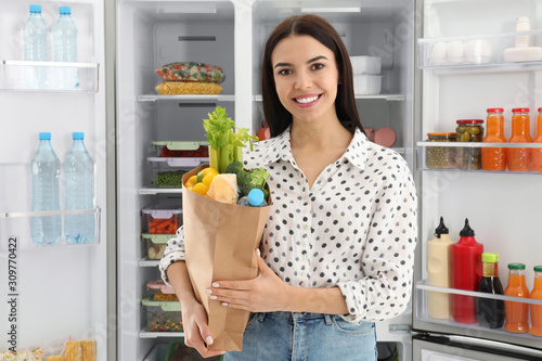 Young woman with bag of groceries near open refrigerator