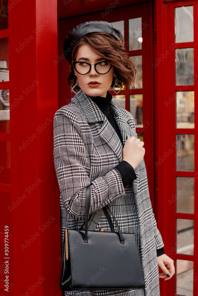 Outdoor portrait of confident fashionable woman wearing classic checkered coat, glasses, beret, holding classic leather handbag, posing near red phone booth