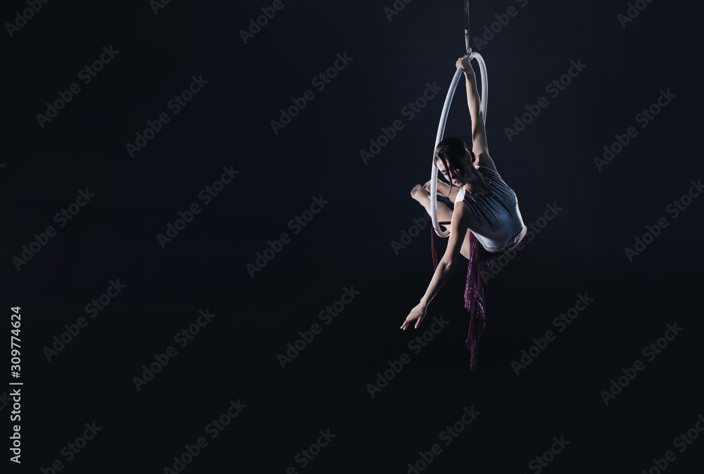 Fototapeta Young woman performing acrobatic element on aerial ring against dark background. Space for text