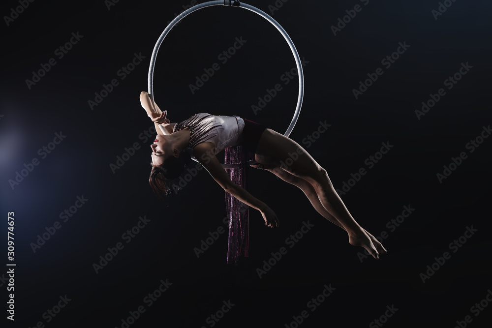Fototapeta Young woman performing acrobatic element on aerial ring against dark background