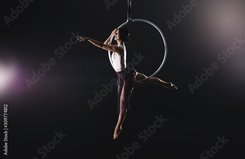 Fototapeta Naklejka Na Ścianę i Meble -  Young woman performing acrobatic element on aerial ring against dark background