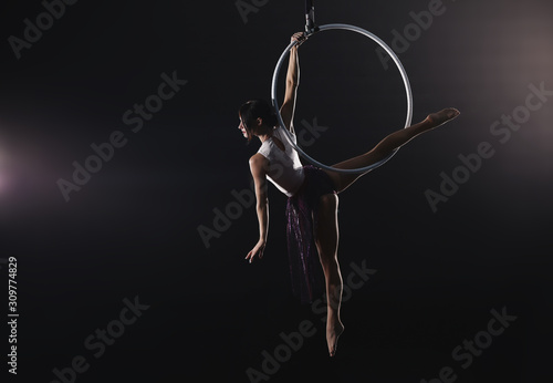 Young woman performing acrobatic element on aerial ring against dark background