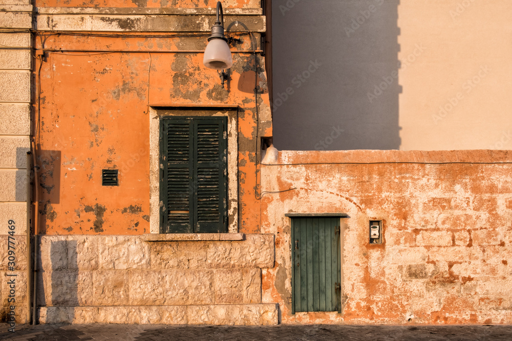 Fototapeta premium Stone wall of the building, morning sun, orange color. Closed window and door, Polignano, Italy.
