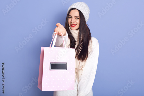 Indoor studio shot of cheerful beautiful female wearing white sweater, hat and scarf, holding paperbag with Christmas present, smiling sincerely, standing isolated over lilac background. Winter time. photo