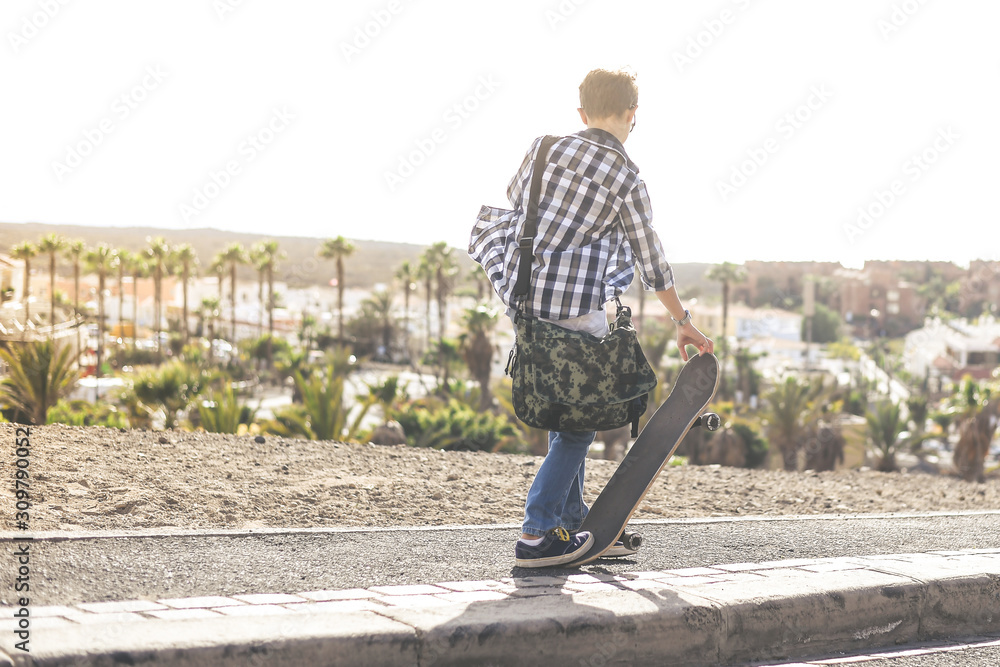 Trendy young boy using skateboard outdoor with bag on the shoulders. Happy teen come back from school with skate, exotic panorama in background. Freedom education carefree youth and lifestyle concept.