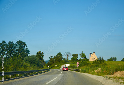 Red car on a road passing a small church