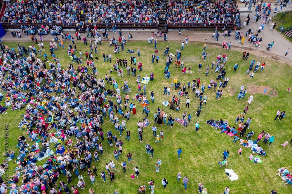 A large crowd of people are placed on the podium, standing and sitting on the grass in front of the stage. Shooting from a drone.