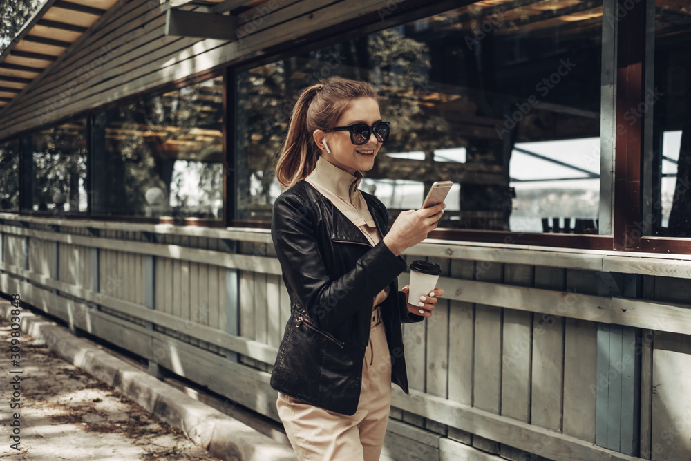 Portrait of Stylish Girl Wears Women Overalls and Black Leather Jacket Drink Coffee, Using Smartphone