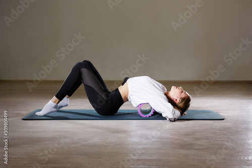 A beautiful slim young girl of European appearance demonstrates exercises with a gymnastic roller for yoga and relaxing her back.