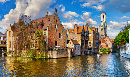 Bruges, Belgium. Ancient medieval european city. View at tower Belfort van Brugge and vintage building at bank of Rozenhoedkaai channel river. Panoramic view with blue sky and clouds. Famous tourist.