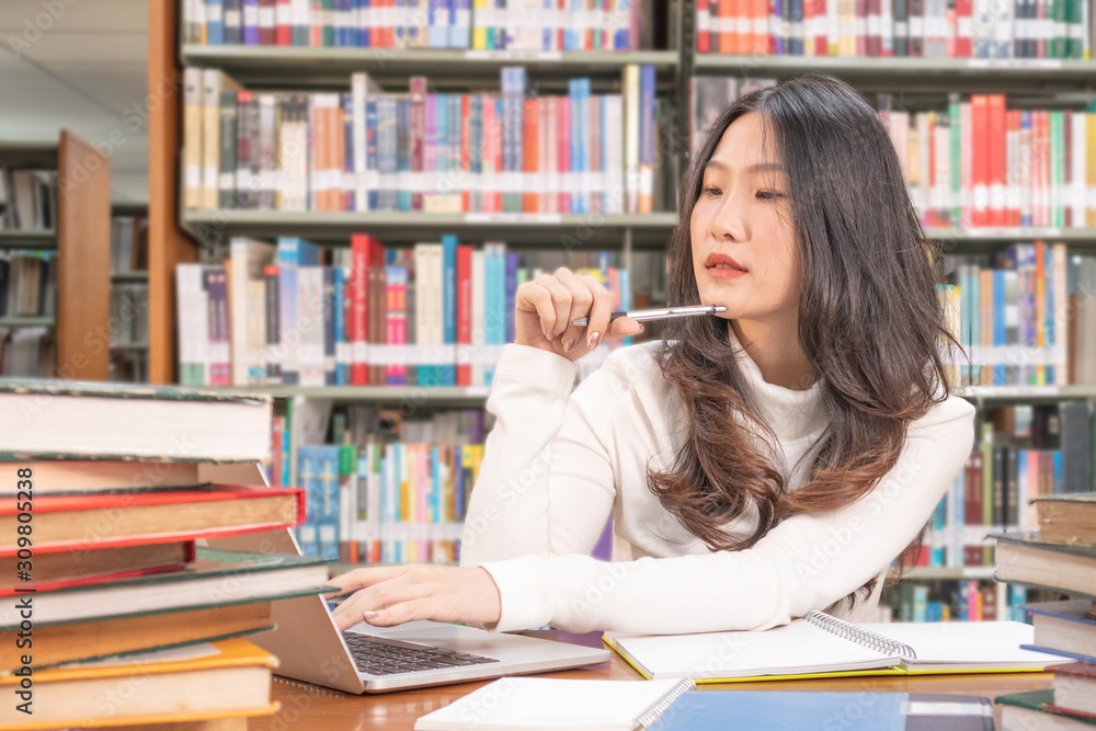 Close up young asian woman sitting and thinking while using laptop on desk in library with bookshelf background before exam university or school, education and searching knowledge concept. Close up young asian woman sitting and thinking while using laptop on desk in library with bookshelf background before exam university or school, education and searching knowledge concept.