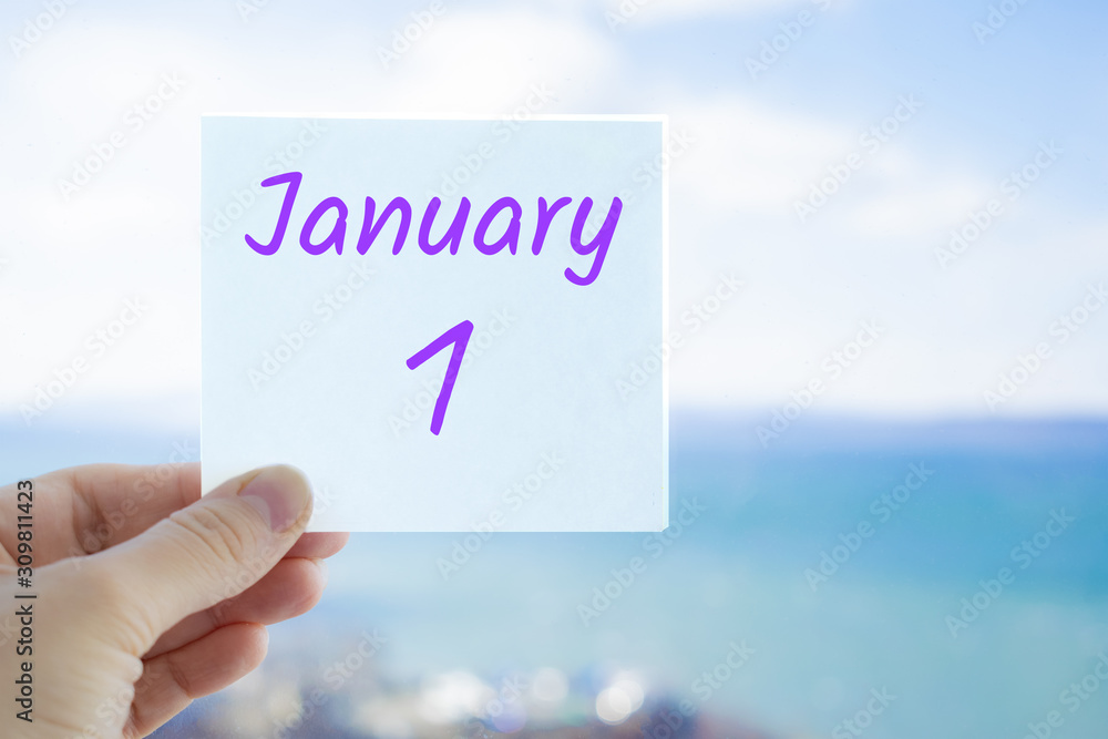January 1st. First day of year. Hand holding sticker with text January 1 on the blurred background of the sea and sky. Copy space for text. Month in calendar concept January 1st. First day of year. Hand holding sticker with text January 1 on the blurred background of the sea and sky. Copy space for text. Month in calendar concept