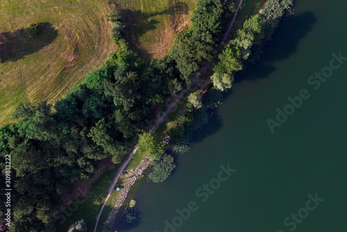 A country path that runs along a lake surrounded by greenery photographed from above by a drone