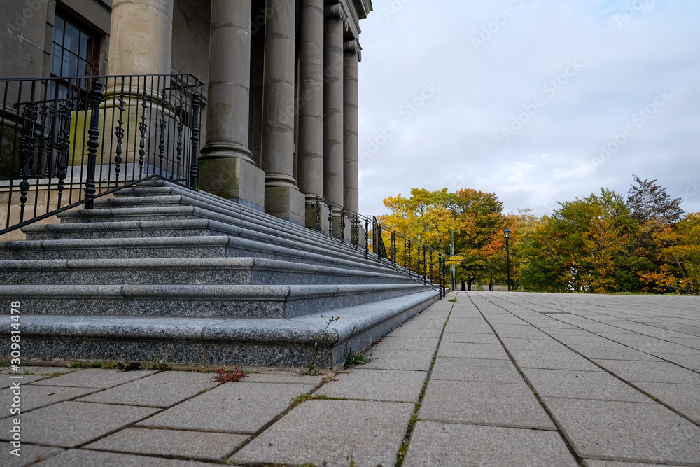 Outside exterior view of a government building. There are marble steps, block steps and columns under a very cloudy sky with trees in the background.