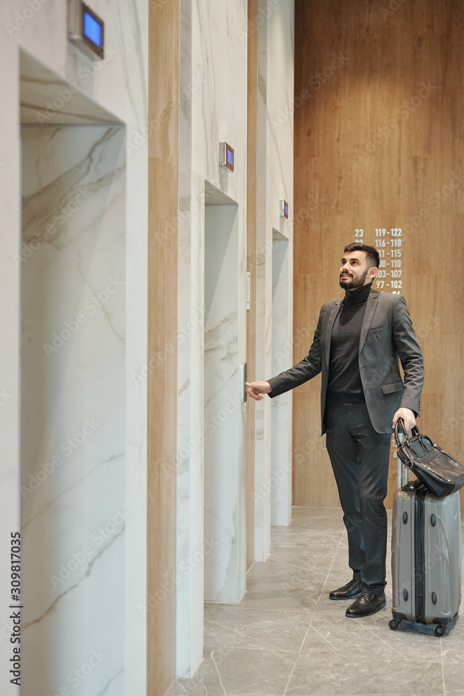 Businessman pushing button on the wall while standing by one of elevator doors Businessman pushing button on the wall while standing by one of elevator doors