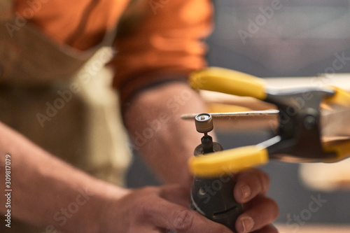 Detail of carpenter's hands filing the edge of a wooden plank with a Dremel tool. There is an out-of-focus clamp holding everything in the foreground.