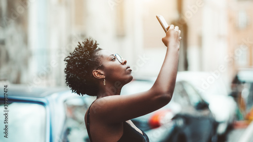 Portrait of an African woman in spectacles taking a photo of a showplace using her smartphone; beautiful young black female in eyeglasses is photographing something above the street via the cellphone