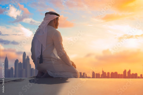 Arabic man with traditional emirates clothes sitting on knees in the UAE desert front Dubai skyline.